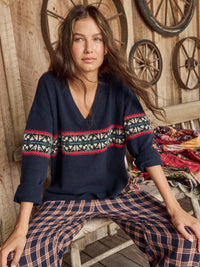 A woman seated on a rustic wooden stool wearing a navy knit sweater with a colorful geometric pattern across the chest and sleeves, paired with navy plaid pants. The background features a wooden wall with decorative wagon wheels.