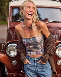 A woman laughing outdoors in front of a vintage brown car, wearing a strapless plaid tube top, blue jeans, and a chunky brown knit cardigan draped off her shoulders.
