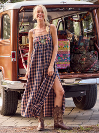 A woman stands in front of the open trunk of a vintage wood-paneled vehicle, wearing a long plaid maxi dress with floral embroidery at the bust and brown cowboy boots.