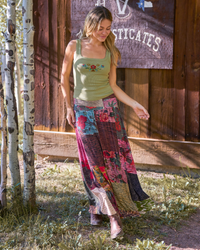 A woman wearing a sleeveless olive green tank top with floral embroidery and a long, colorful patchwork maxi skirt stands outdoors in front of a rustic wooden wall and birch trees.