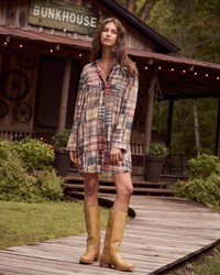 A woman stands on a wooden boardwalk in front of a rustic cabin with a porch and string lights, wearing a long-sleeve plaid shirt dress in beige, red, and blue tones, paired with tan cowboy boots.