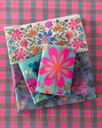 Three folded fabric sheets with colorful floral patterns are stacked on top of each other. The sheets feature vibrant designs including pink, blue, yellow, and orange flowers on a light background, set against a pink and blue gingham checkered surface.