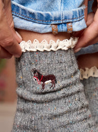 Close-up of a person wearing a gray knit sock with a brown embroidered donkey and red harness detail, paired with a white lace trim and blue denim pants.